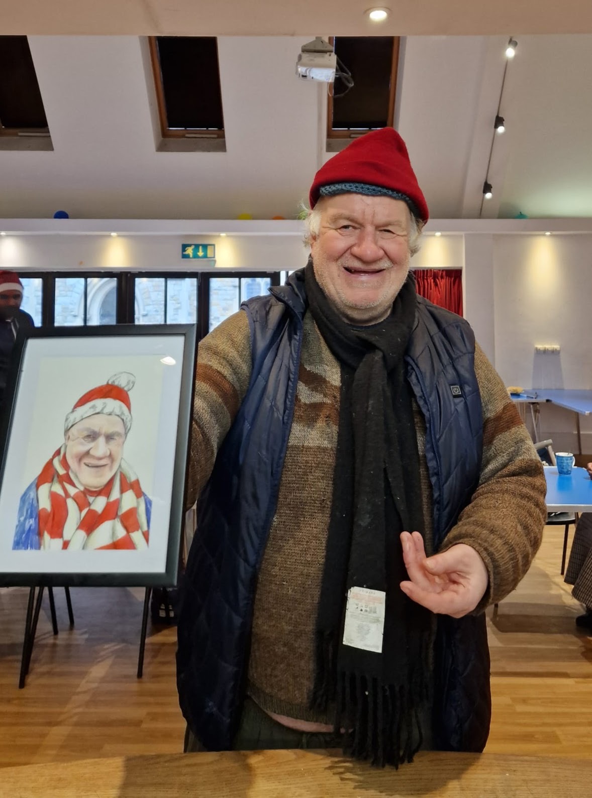 An older man wearing a red beanie, scarf, and winter clothes smiles while holding a framed portrait of himself dressed similarly. He stands in a bright room with tables and chairs in the background.