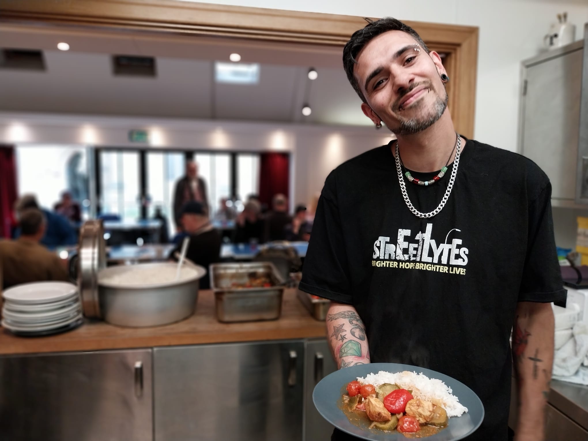 A smiling person holds a plate of rice and curry in a kitchen, with pots of food and stacked plates on the counter. People are seated and talking in the background.