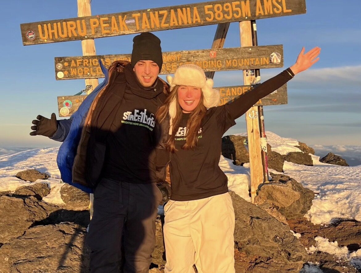 Two people dressed in winter clothing stand smiling with arms around each other at Uhuru Peak, Tanzania, 5895 meters above sea level, with a snowy landscape and signposts in the background.
